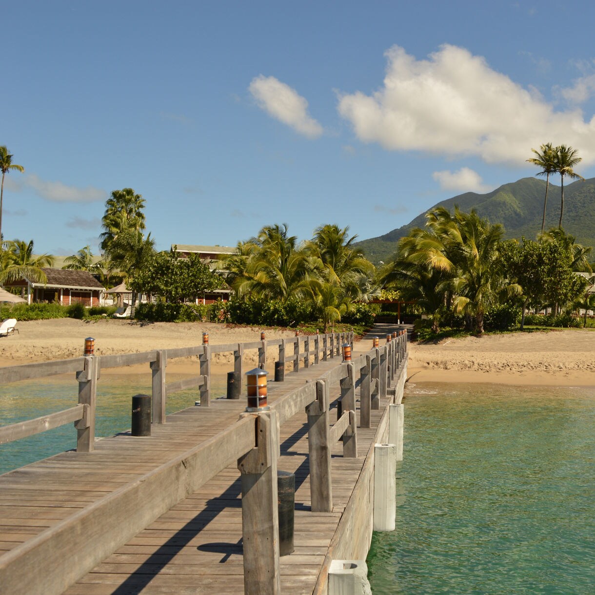 A wooden pier stretches out into calm blue waters, framed by palm trees and a view of St. Kitts' verdant hills and clear skies.