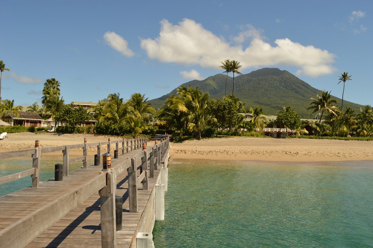 A wooden pier stretches out into calm blue waters, framed by palm trees and a view of St. Kitts' verdant hills and clear skies.