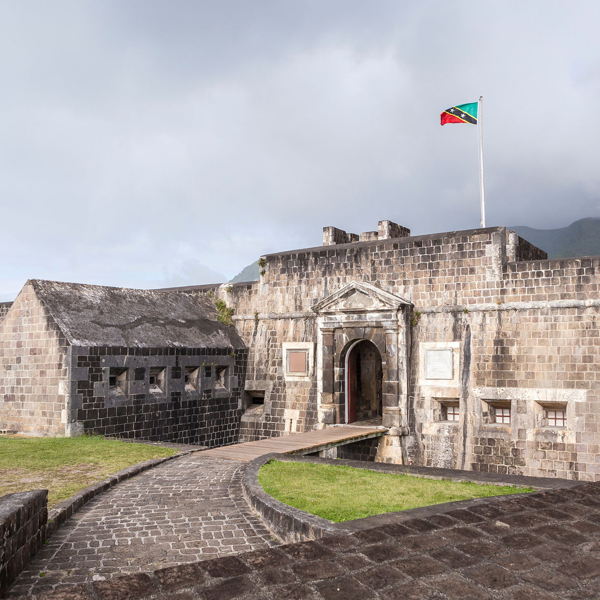 The entrance to Brimstone Hill Fortress in St. Kitts, featuring the stone archway, the national flag flying above and the surrounding lush hills.