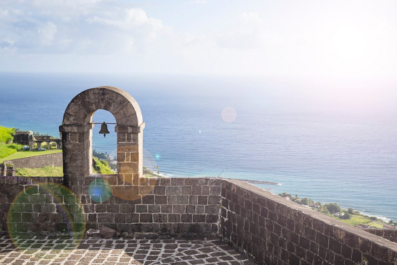 A bell tower at Brimstone Hill Fortress on St. Kitts stands against the backdrop of the ocean, surrounded by lush greenery and clear skies.