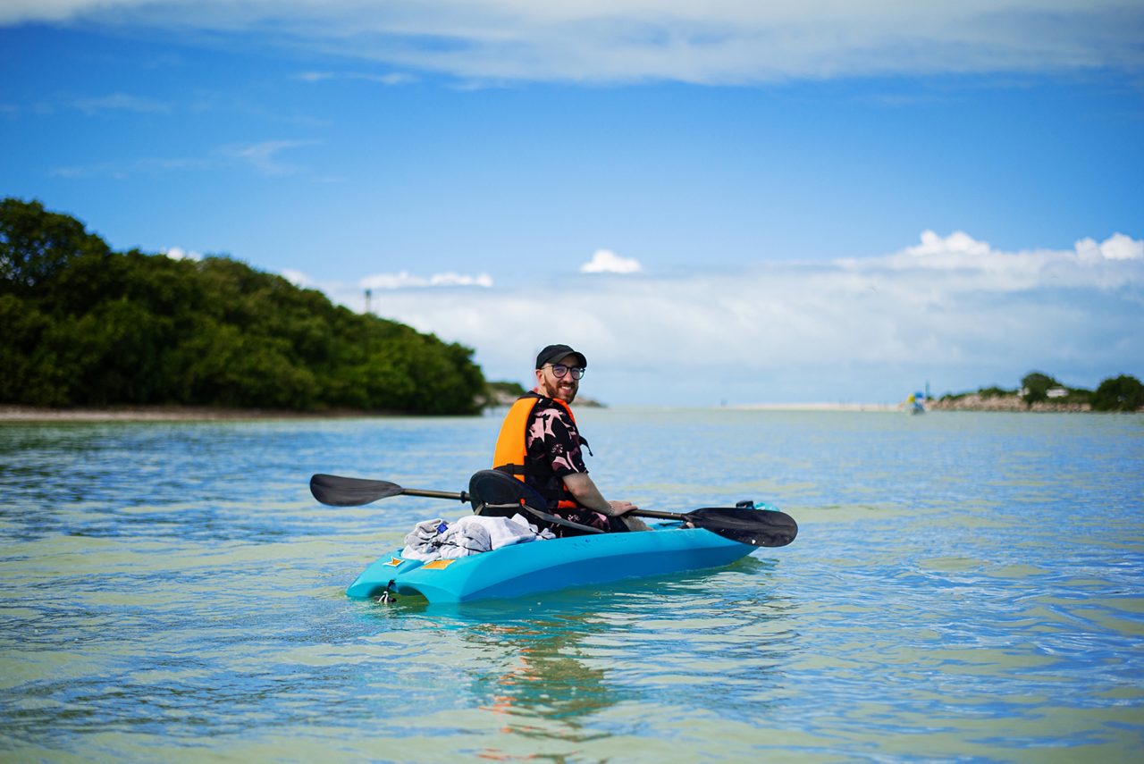 A person kayaking in the calm waters off the coast of St. Kitts, surrounded by green hills and a clear blue sky.