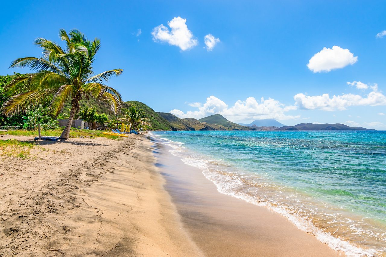 A serene view of Cockleshell Beach in St. Kitts, with clear blue water, golden sand and palm trees under a sunny sky, leading to a distant mountain range.