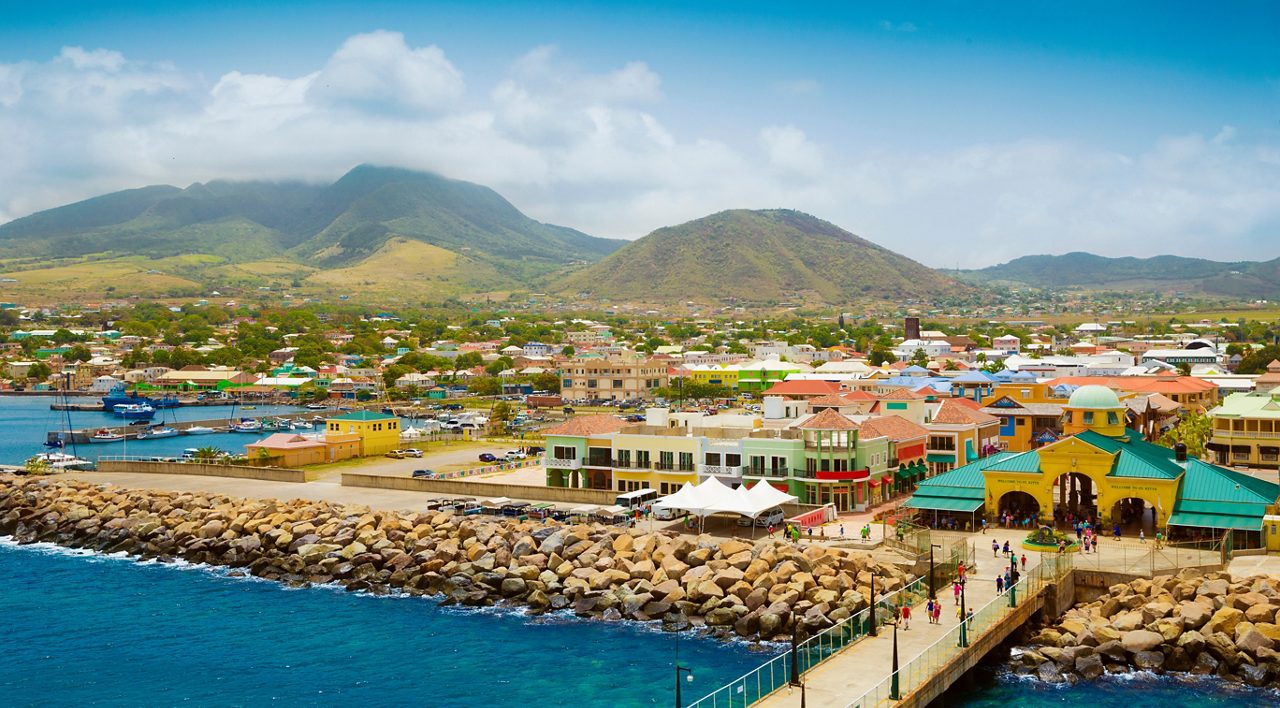A scenic view of Basseterre, St. Kitts, featuring a bustling waterfront, pastel-colored colonial buildings, a cruise port entrance with a green-roofed pavilion, and towering mountains in the background.