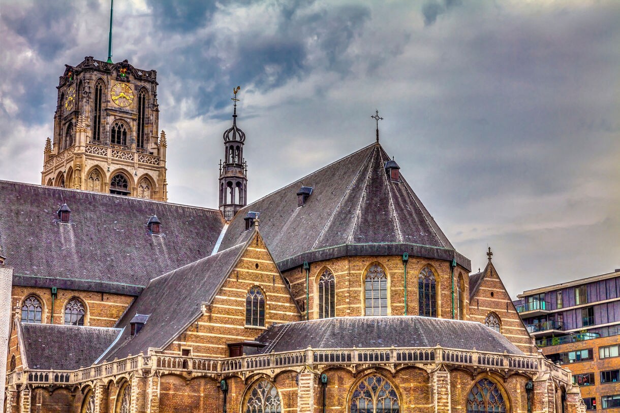 St. Laurenskerk in Rotterdam with its ornate Gothic tower and arched windows, framed against a cloudy sky with modern buildings nearby.