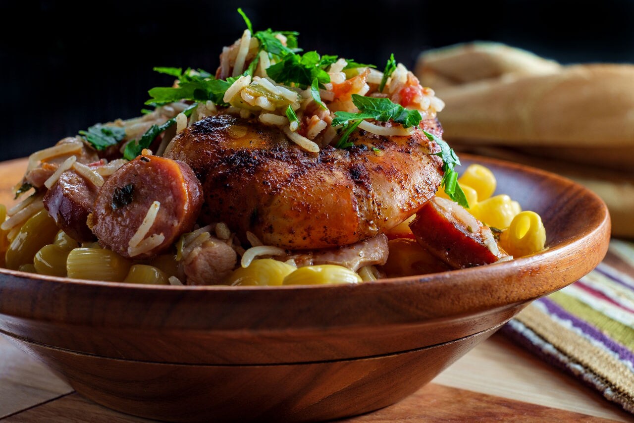 A close-up of a traditional St. Lucian Creole dish featuring grilled chicken, sausage, rice and pasta, garnished with fresh herbs in a wooden bowl.