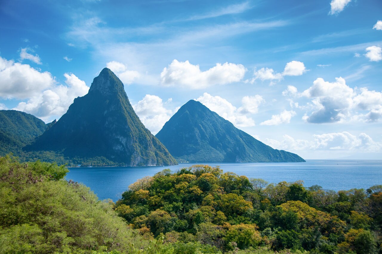 The towering Pitons of St. Lucia covered in green vegetation, set against a bright blue sky with scattered clouds, overlooking the deep blue waters of the Caribbean Sea.