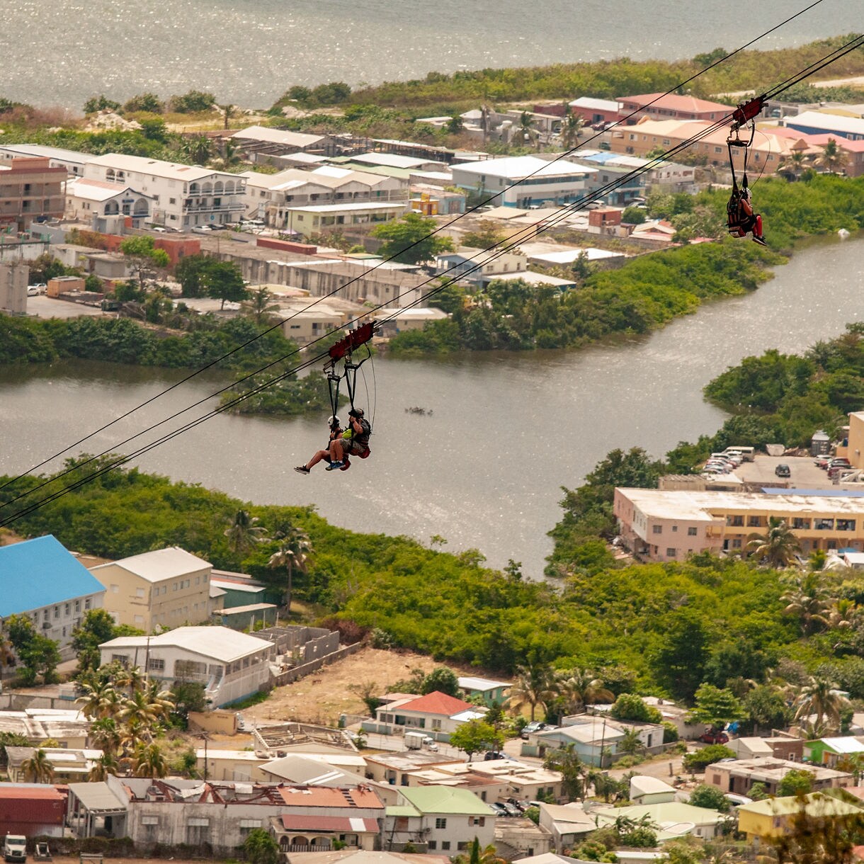 Two people ziplining over St. Maarten’s landscape, with a view of the city and a winding river below.