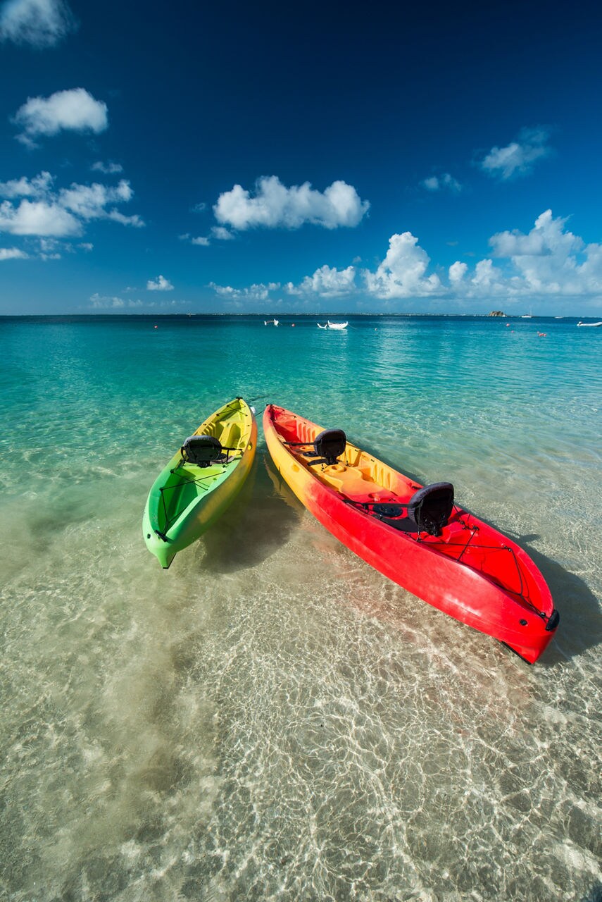 Two colorful kayaks, green and red, are sitting in shallow, clear waters with a vibrant sky above and distant boats in the background.