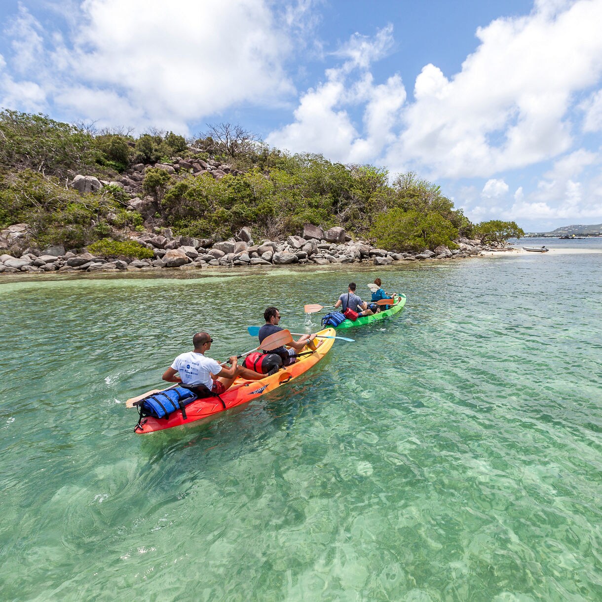 Four people kayak through a calm lagoon in St. Maarten, passing rocky terrain and vibrant greenery under a bright blue sky