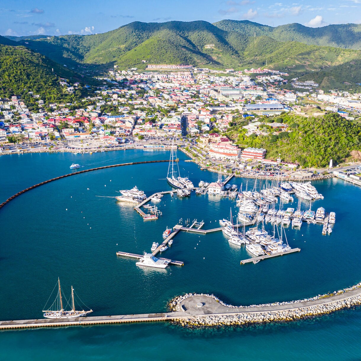A circular marina filled with yachts and sailboats sits in a calm blue bay, surrounded by a protective breakwater