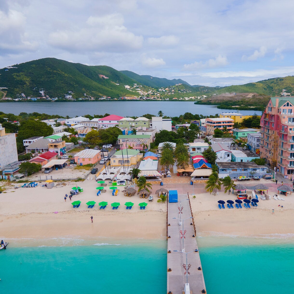 An aerial view of St. Maarten’s beach with colorful buildings and green and blue umbrellas scattered across the sand, leading to a pier over turquoise waters. The surrounding mountains complete the picturesque scene.
