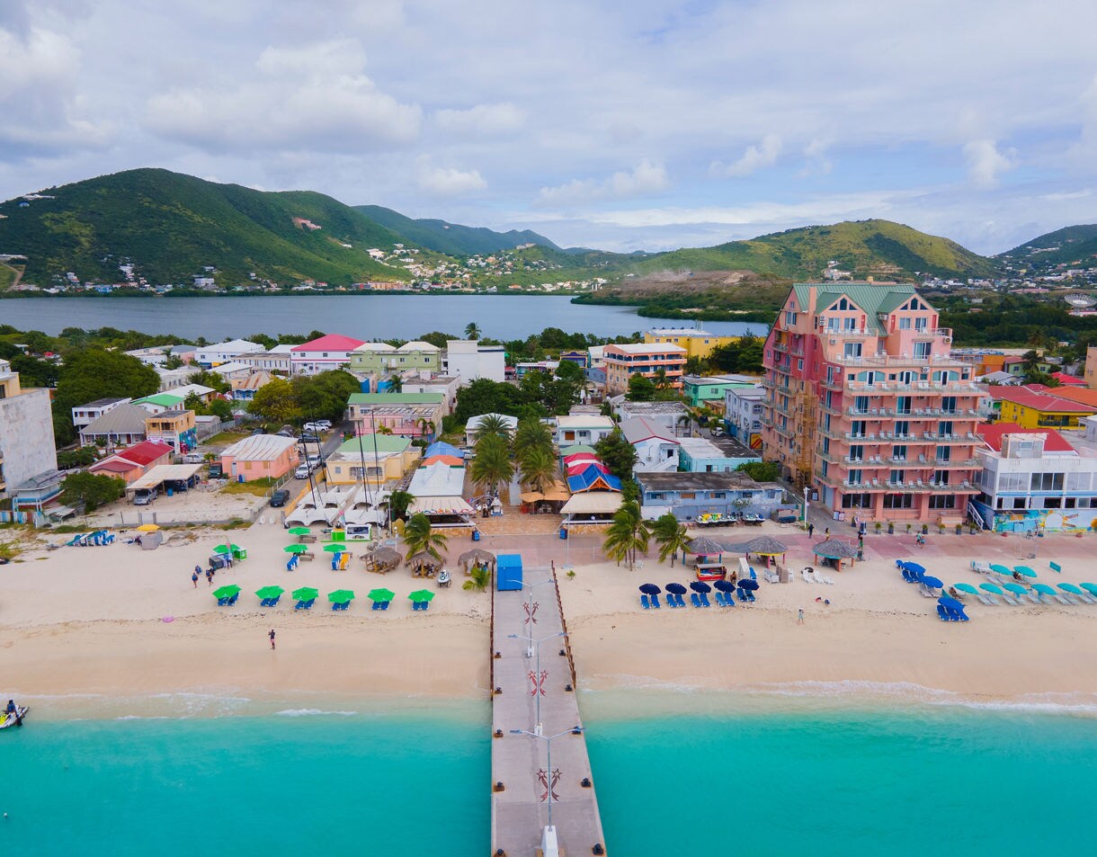 An aerial view of St. Maarten’s beach with colorful buildings and green and blue umbrellas scattered across the sand, leading to a pier over turquoise waters. The surrounding mountains complete the picturesque scene.