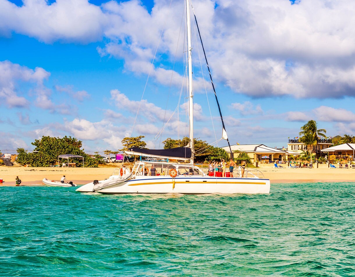 A catamaran with passengers aboard cruises through crystal-clear water, with the sandy beach and colorful beachside buildings of St. Maarten in the background under a sunny sky.