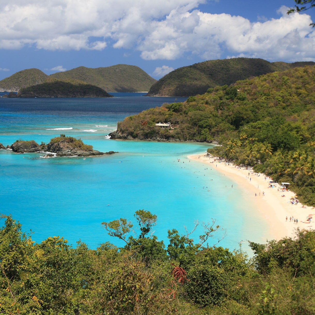 Elevated view of a crescent shaped beach with bright turquoise water, small rocky islets, lush green hills and people relaxing along the sandy shoreline.