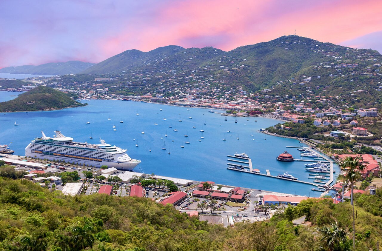A panoramic view from Paradise Point in St. Thomas, showing a cruise ship docked at the harbor, with the town's colorful buildings and hills in the background. The sky features a warm, pinkish hue as the sun sets.