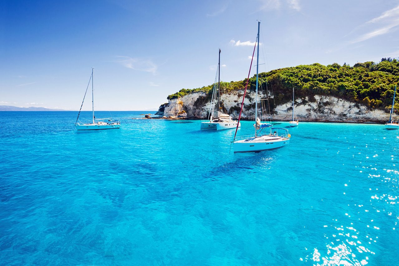 Several sailboats anchored in the vibrant turquoise waters of St. Thomas, with lush green cliffs and white limestone formations in the background under a clear blue sky.