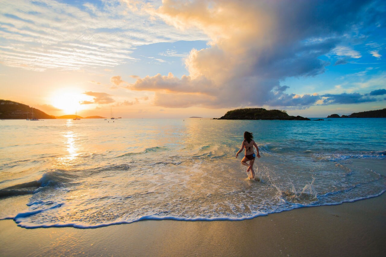 A person running in the shallow surf at a St. Thomas beach during sunset, with the sun reflecting off the water and boats in the distance. The sky features soft clouds, creating a peaceful and scenic atmosphere.