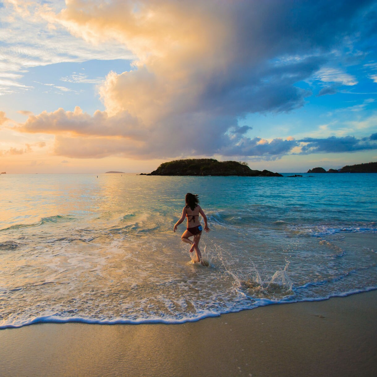 A traveler splashing into shallow ocean water at sunset on a Caribbean beach, with glowing skies, rolling waves and small islands stretching across the horizon.