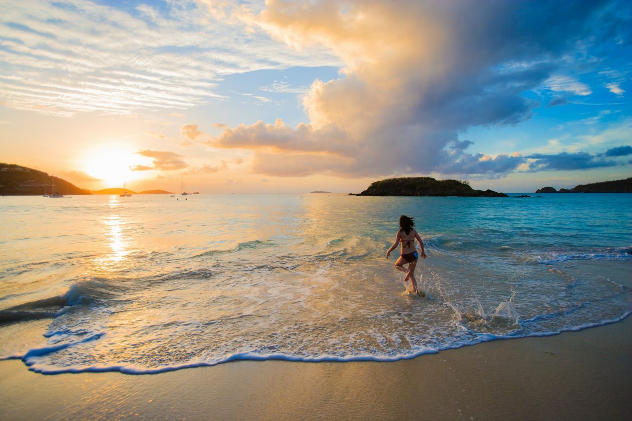 A person running in the shallow surf at a St. Thomas beach during sunset, with the sun reflecting off the water and boats in the distance. The sky features soft clouds, creating a peaceful and scenic atmosphere.