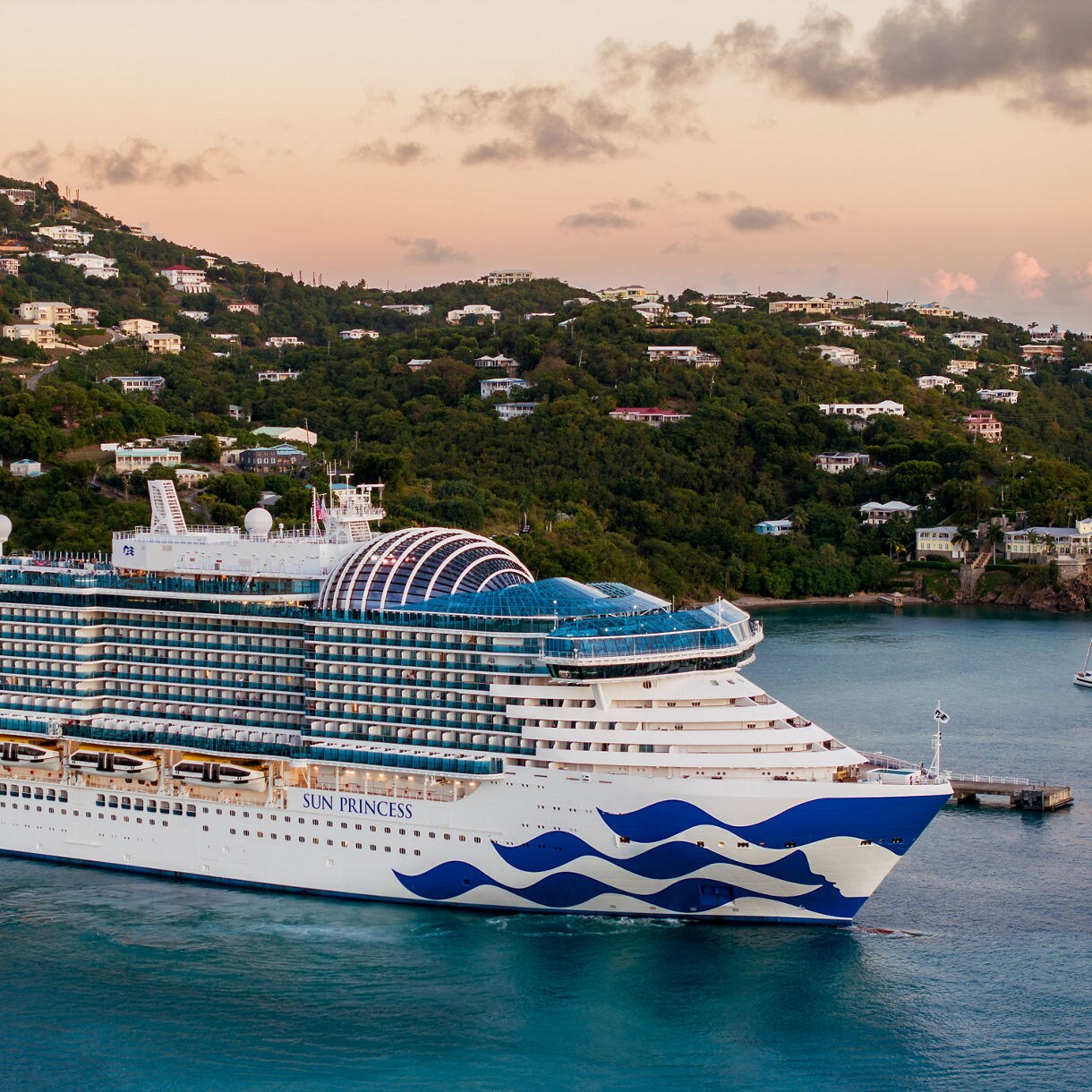 The image shows a brightly lit cruise ship captured from above as it moves through dark waters at night. Vivid blue and white waves trail behind the vessel, creating a striking visual contrast. The ship's deck is illuminated with warm lights, and a swimming pool is visible at the center. The overall scene conveys a sense of luxury, travel, and nighttime adventure.
