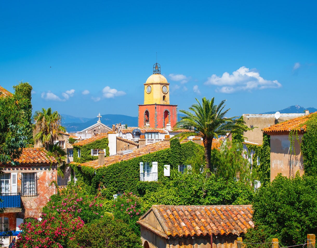 View of Saint-Tropez with terracotta rooftops, lush greenery and a yellow-and-red bell tower rising under a clear blue sky.