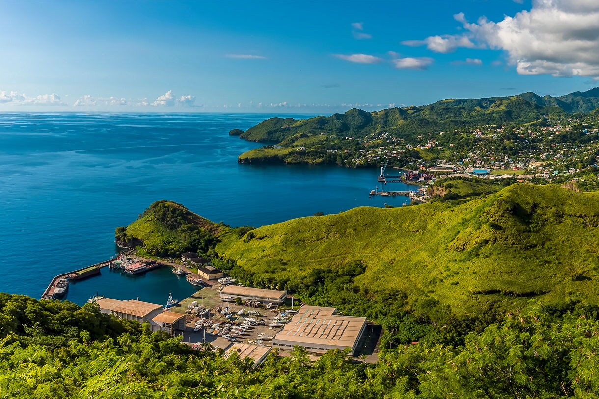 Aerial view of St. Vincent featuring lush green hills, a coastal town with colorful buildings, a small harbor with boats and deep blue ocean waters stretching to the horizon.