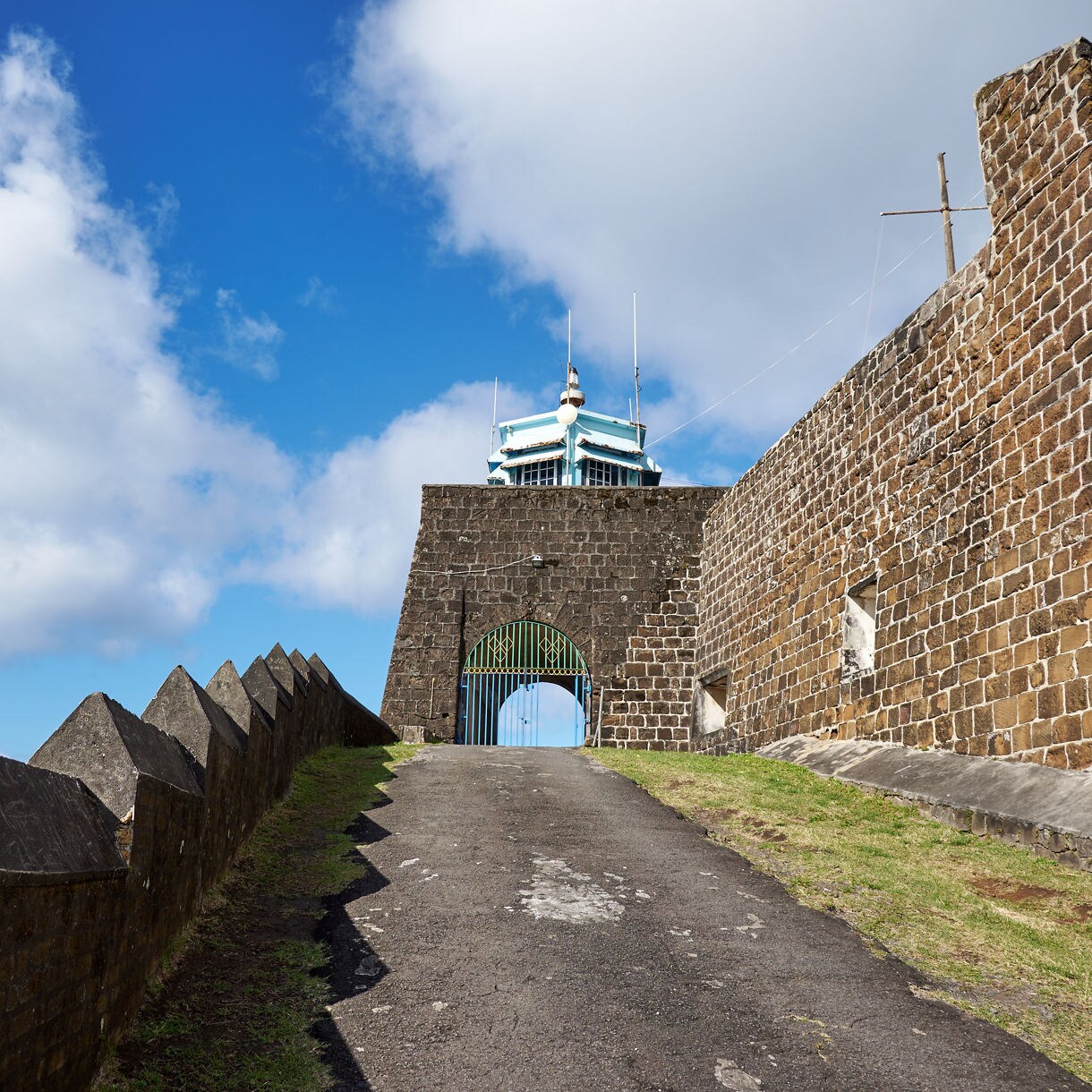 Stone pathway leading to Brimstone Hill Fortress entrance with fortress walls and lighthouse visible against blue sky.