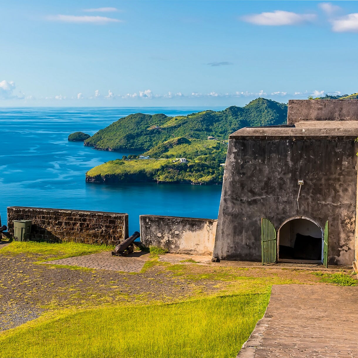Historic cannons line the stone walls of Fort Charlotte in St. Vincent, overlooking a bright blue sea and a lush green peninsula under a clear sky