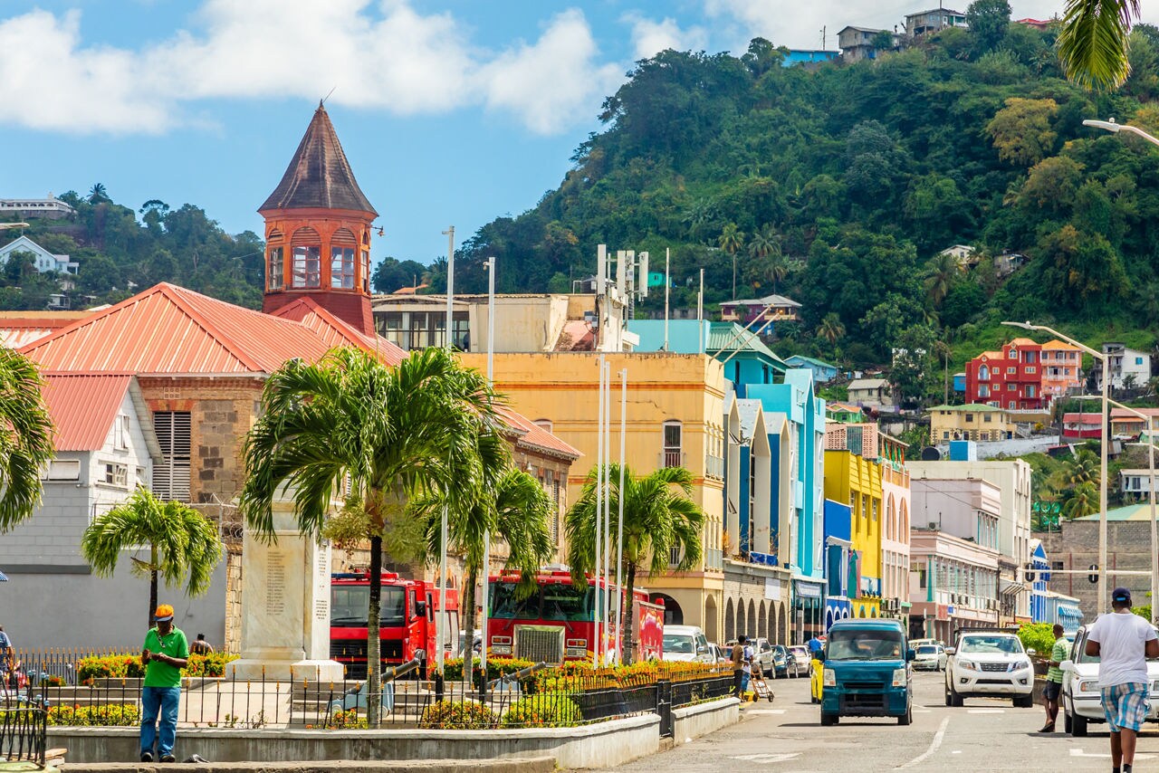 Bustling street in Kingstown, St. Vincent lined with palm trees, colorful colonial-style buildings and a red clock tower, with green hills and homes in the background.