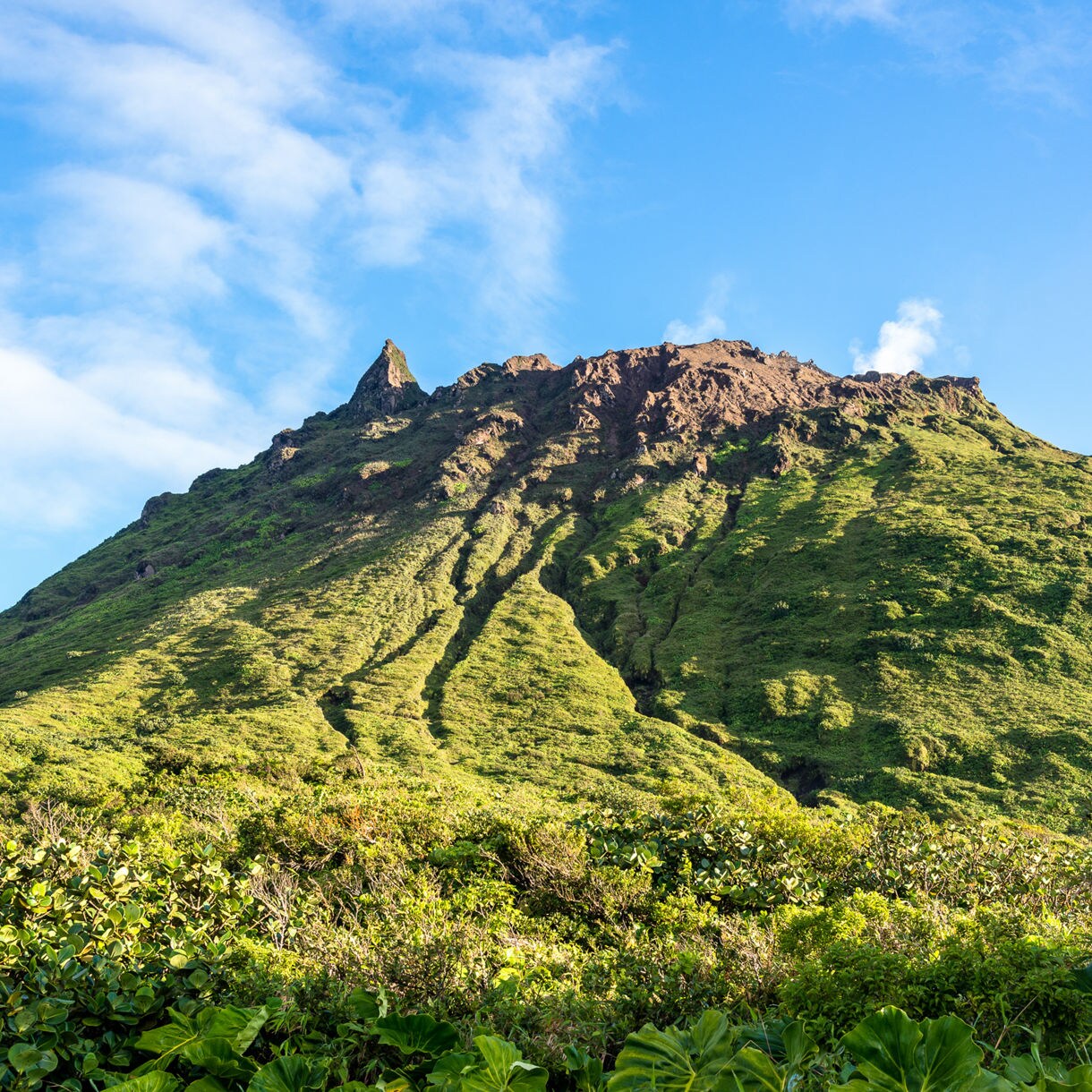 La Soufrière volcano in St. Vincent covered in lush green vegetation with rocky ridges and a small plume of steam rising near the summit under a blue sky