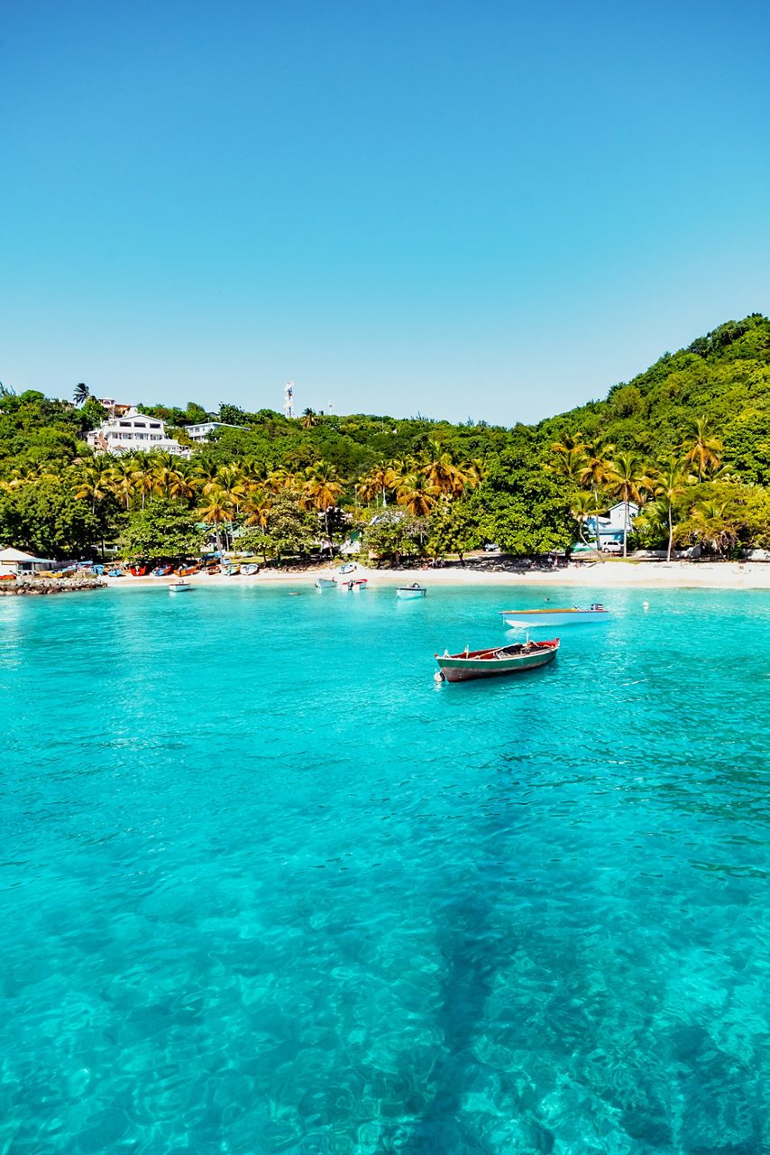Small boats floating in the clear, shallow waters of Britannia Bay off Mustique Island, with a palm-lined shoreline and forested hillside in the background.