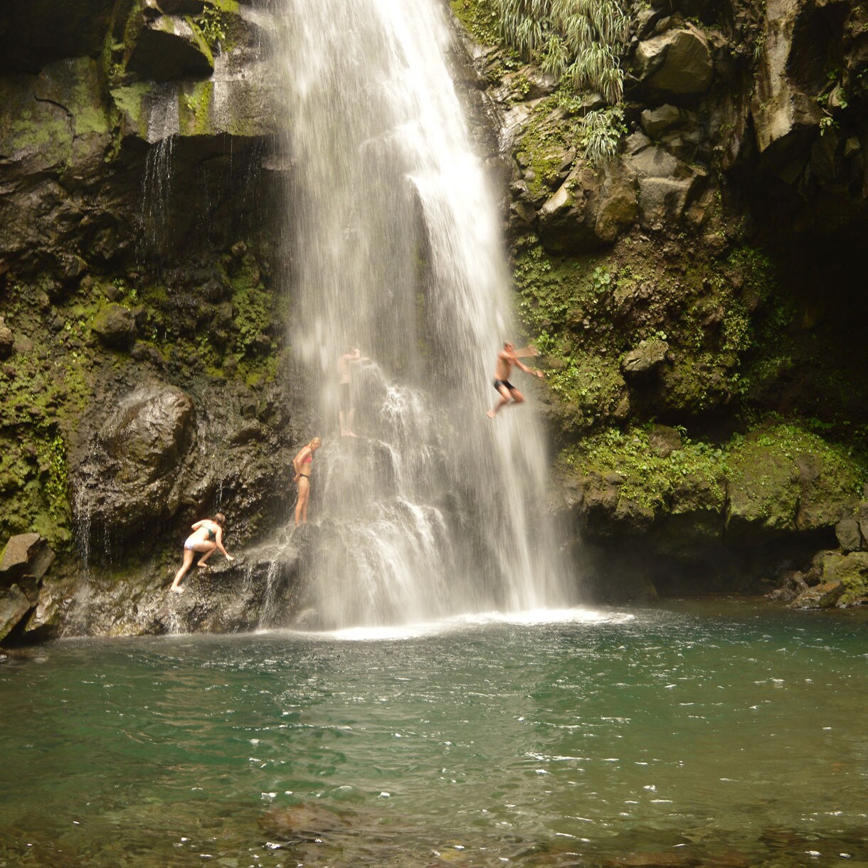 People climbing and jumping off rocks under a cascading jungle waterfall into a clear pool surrounded by mossy cliffs and greenery in St. Vincent