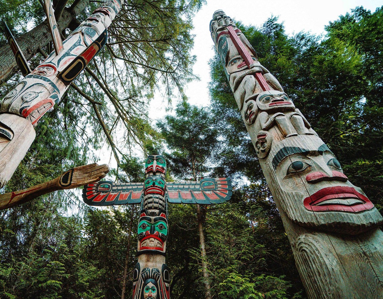 Upward view of several colorful Indigenous totem poles surrounded by tall trees in Stanley Park.