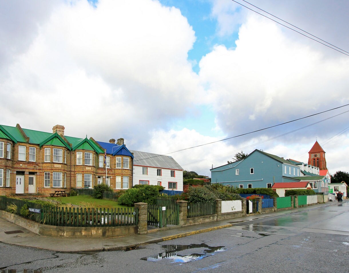Row of brick townhouses with green and blue roofs along a wet street in Stanley, Falkland Islands, with picket fences, scattered homes and a church tower in the background.