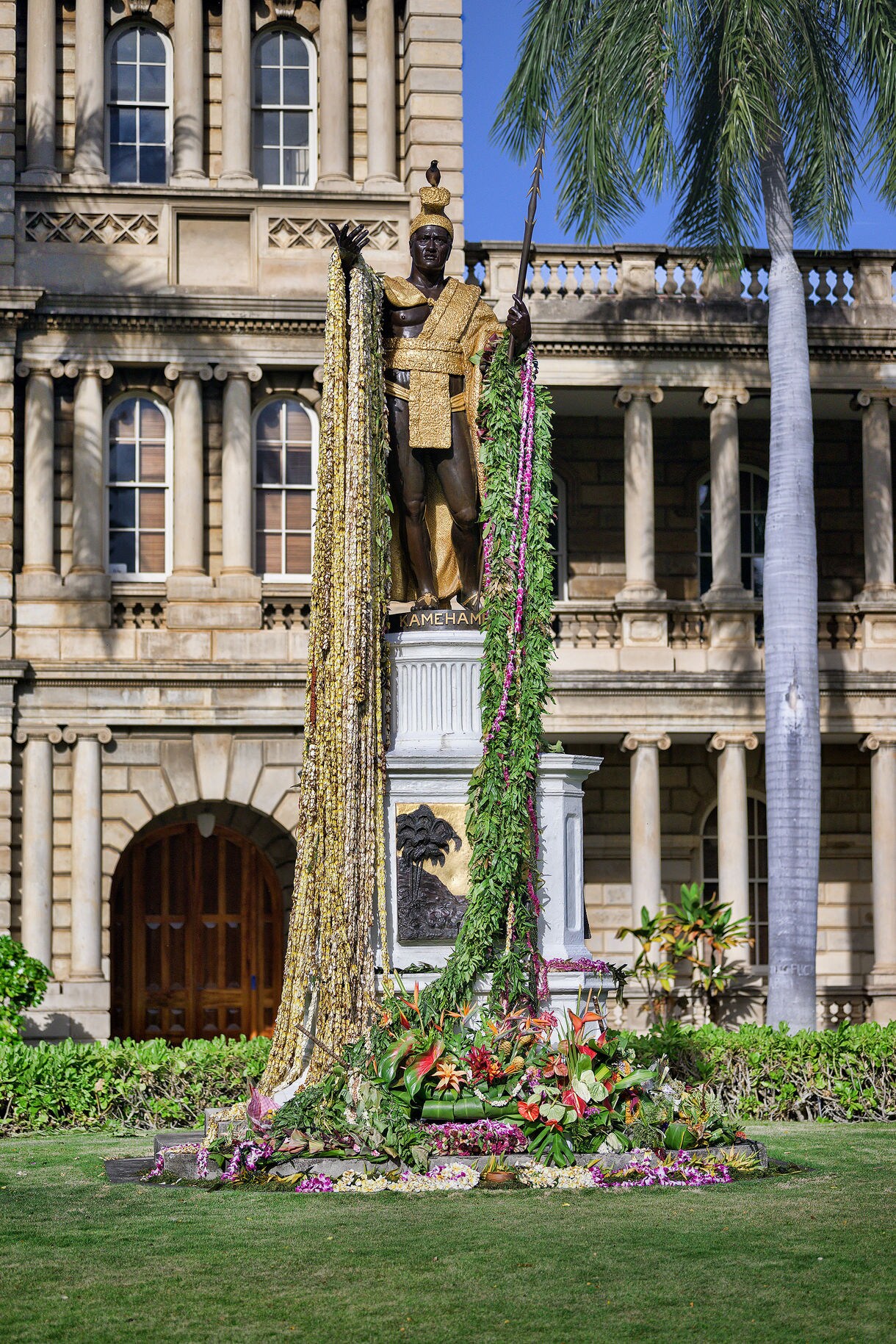 Bronze statue of King Kamehameha in golden regalia, draped with flower leis, in front of the historic Aliʻiōlani Hale building in Honolulu.
