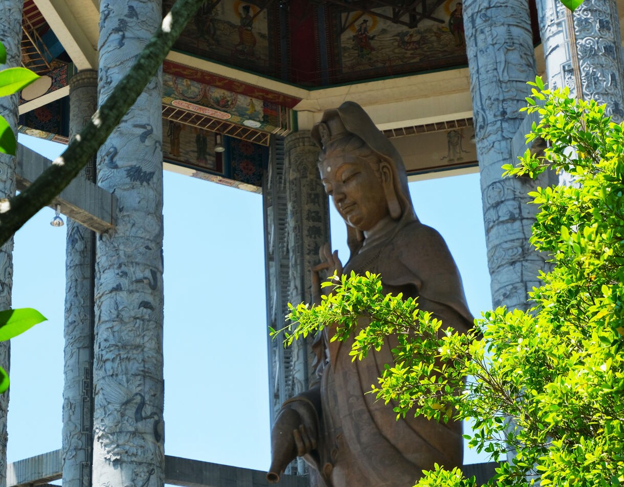  A large Guanyin statue stands under an ornate pavilion with carved stone pillars, partially framed by green foliage and blue sky.