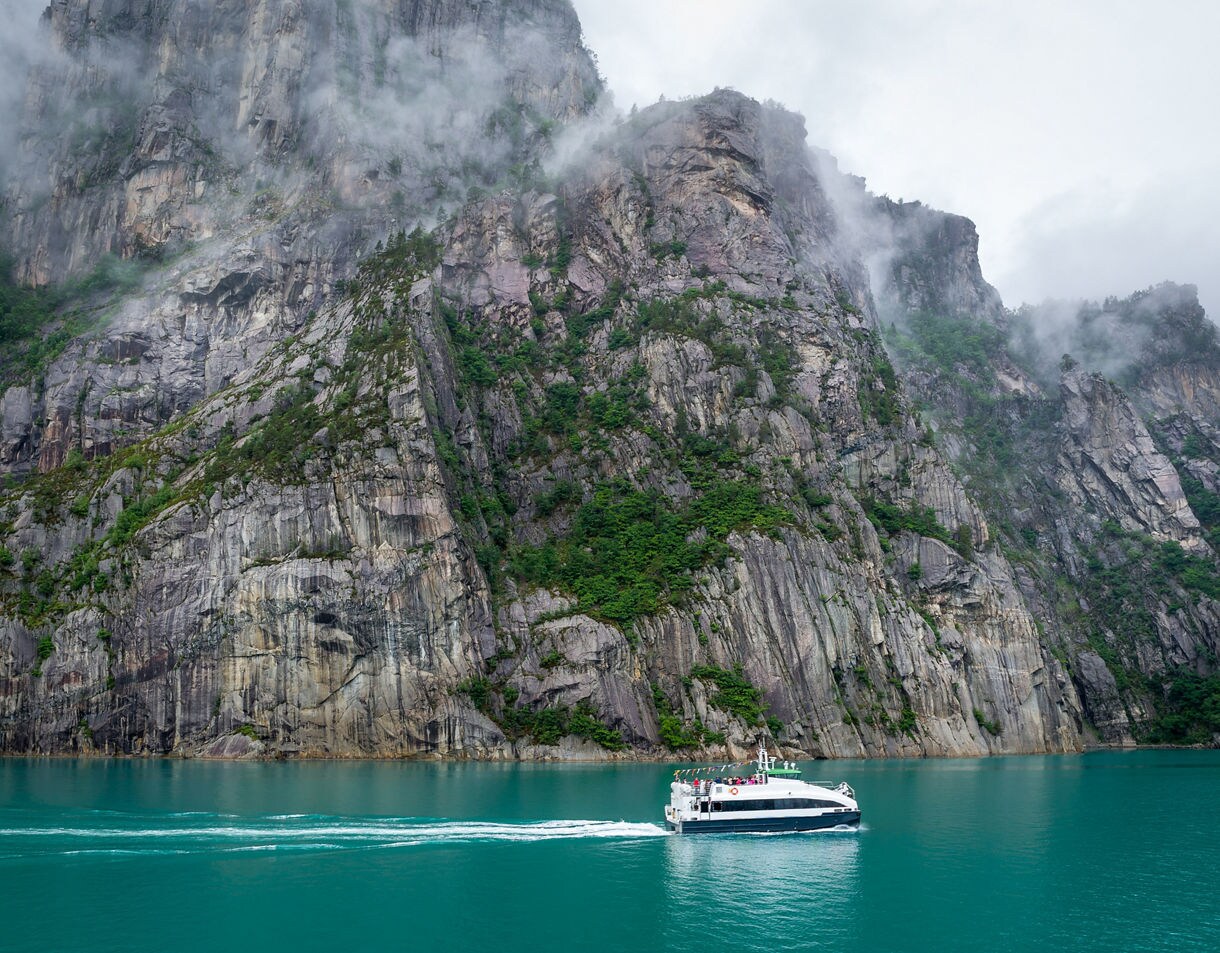 A tour boat sailing on calm turquoise water beside steep rocky cliffs covered in patches of green vegetation and low-hanging mist.