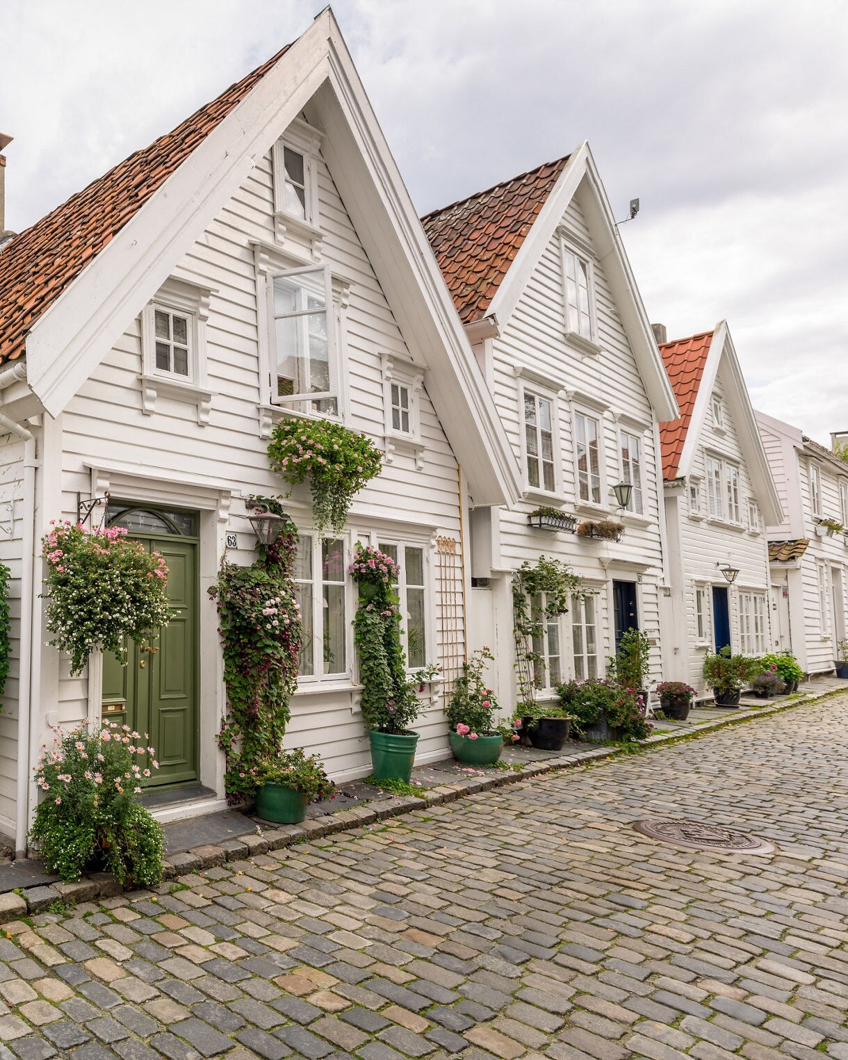 A cobblestone street with white wooden houses featuring red-tiled roofs and potted flowers, creating a cozy historic neighborhood scene.