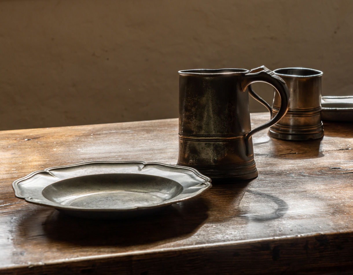 Two metal mugs and two pewter plates arranged on a worn wooden table lit by soft natural light.