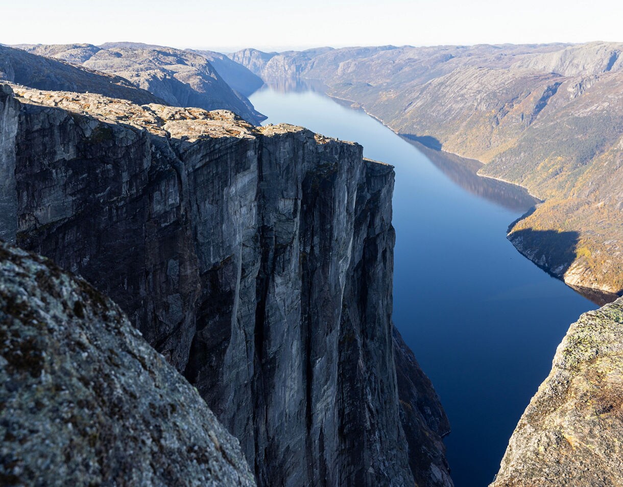 A dramatic cliff rising high above a long, narrow blue fjord, with steep rocky mountains stretching into the distance under clear daylight.