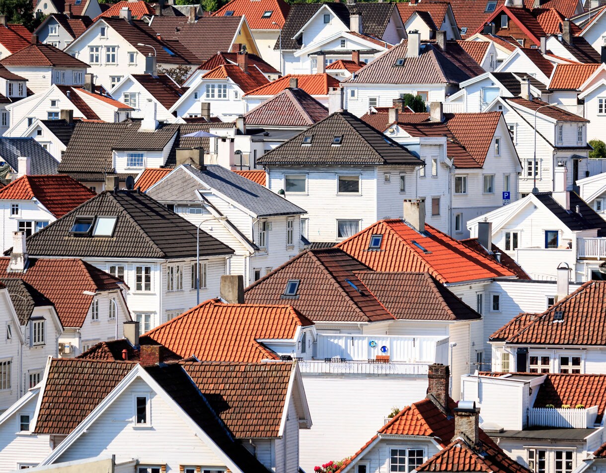 A dense cluster of white wooden houses with red and brown tiled roofs, closely packed together on a sloping neighborhood under daylight.