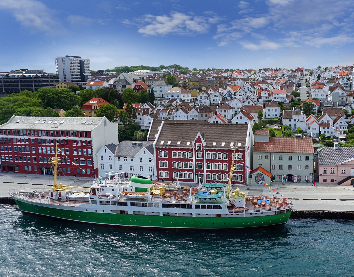 A green and white ship docked along a waterfront lined with pastel and red buildings, with rows of white houses climbing a hillside under a bright blue sky.
