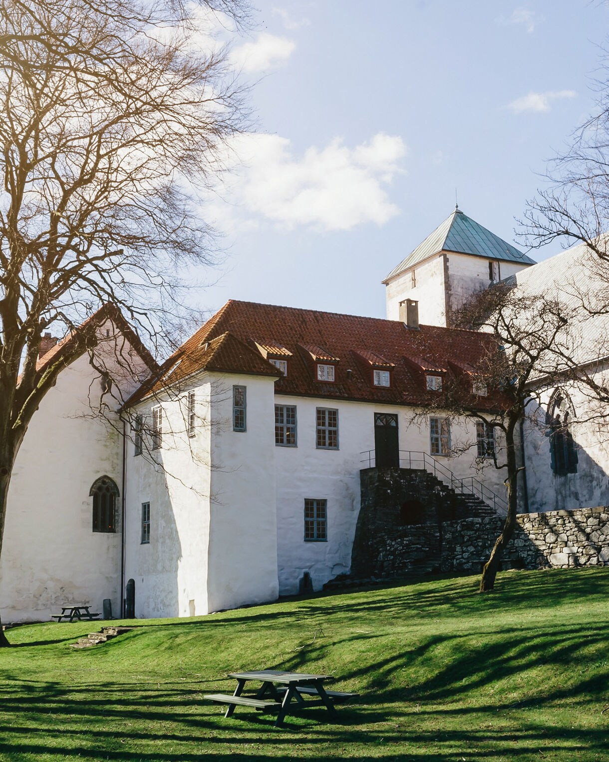 A white stone monastery with red-tiled roofs and a central tower, set on a grassy hillside with leafless trees casting long shadows in the sunlight.