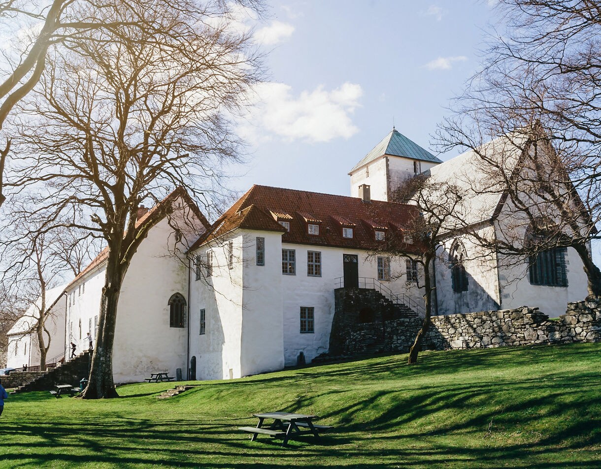 A white stone monastery with red-tiled roofs and a central tower, set on a grassy hillside with leafless trees casting long shadows in the sunlight.