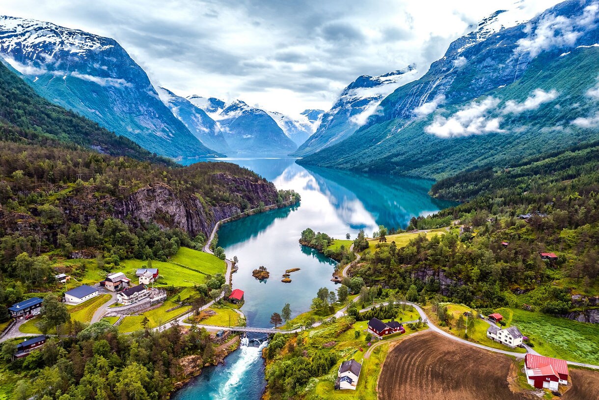 A turquoise fjord winding between steep green and snow-topped mountains, with small farms and houses scattered along the shoreline and a waterfall flowing into the water.
