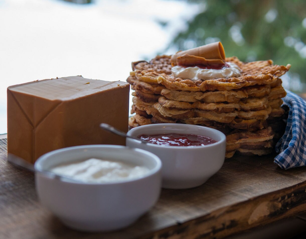 A stack of waffles served with a block of brown cheese, a bowl of cream and a bowl of red jam on a wooden board.