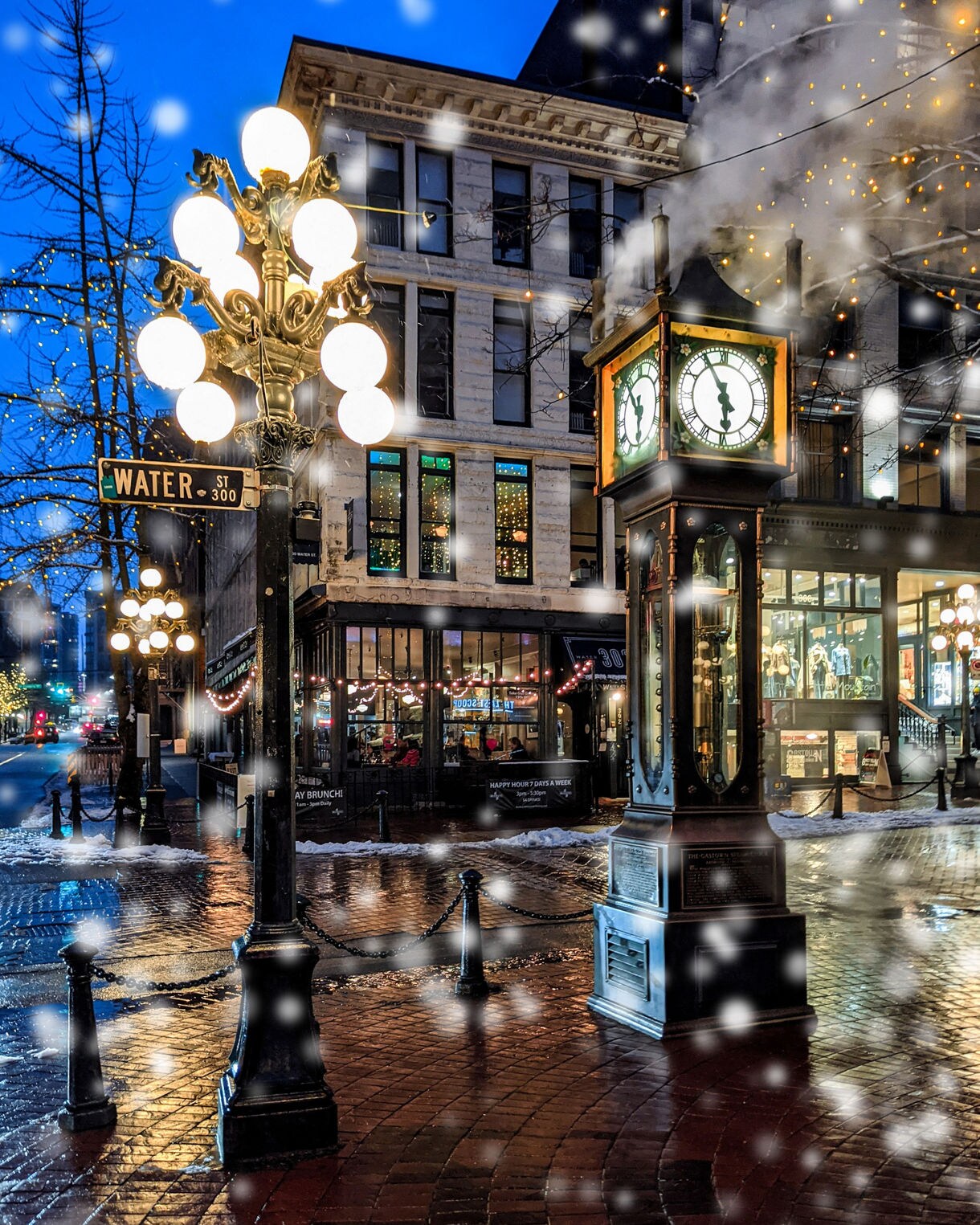 The illuminated Gastown steam clock and lamppost on Water Street at dusk, with snow gently falling over wet cobblestones.