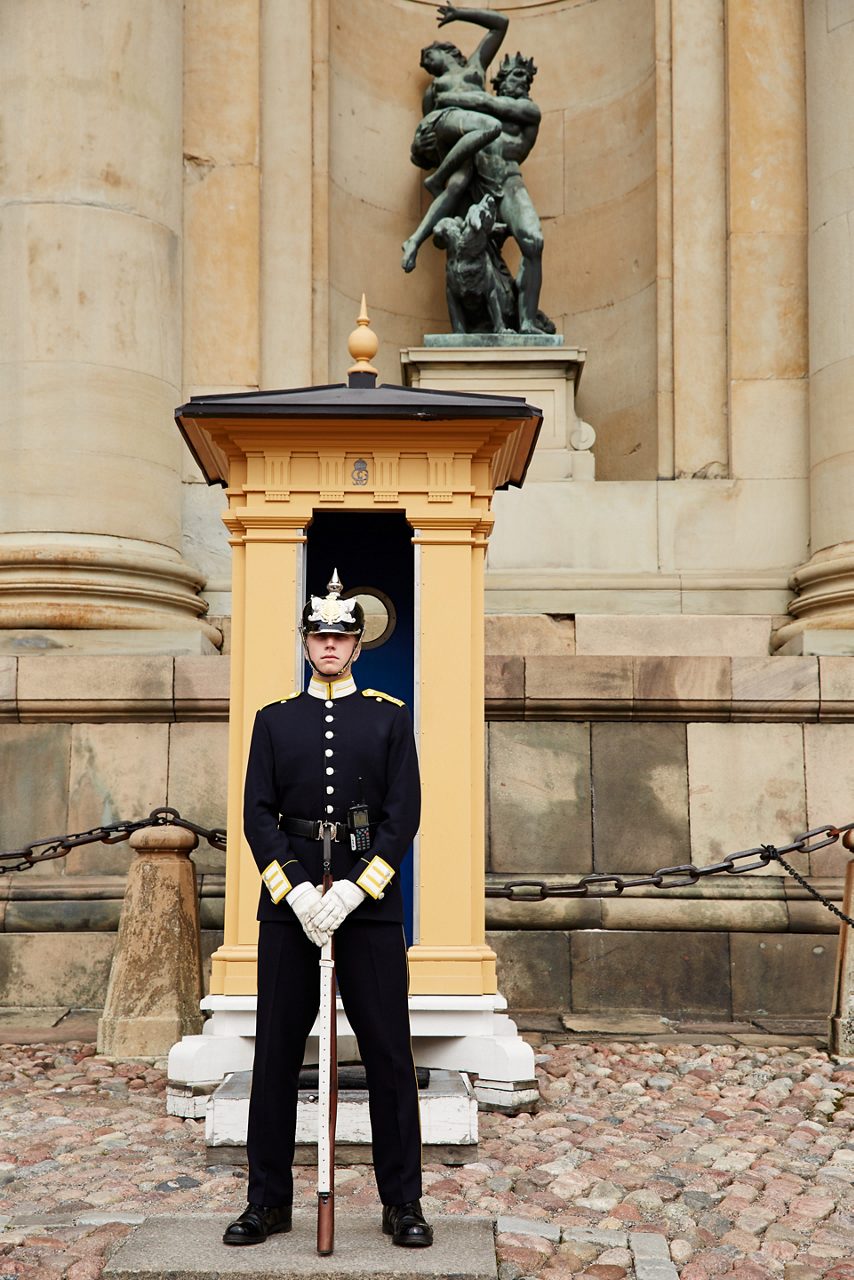 A palace guard in traditional uniform stands watch outside the Royal Palace in Stockholm, Sweden, with the historic architecture of the palace in the background. 