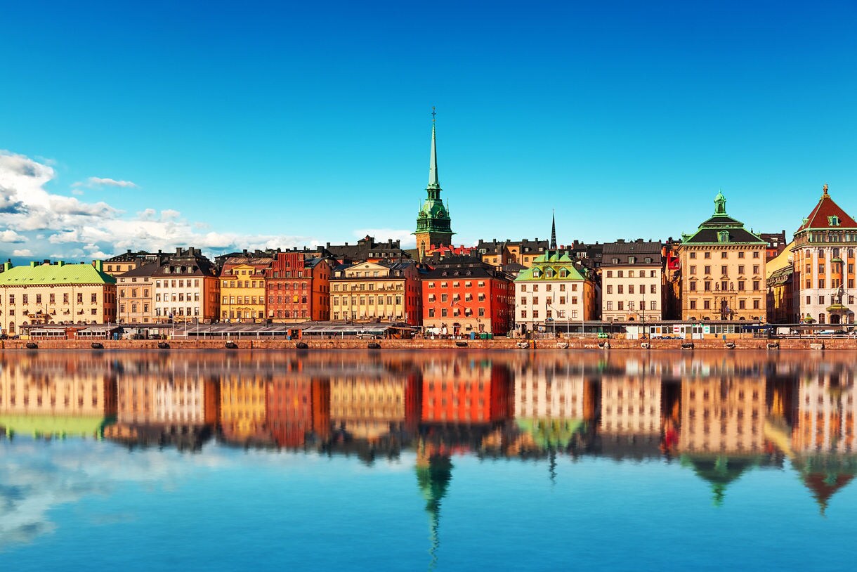 The vibrant skyline of Stockholm's Gamla Stan, with colorful historic buildings reflected in the calm water on a bright, clear day. 