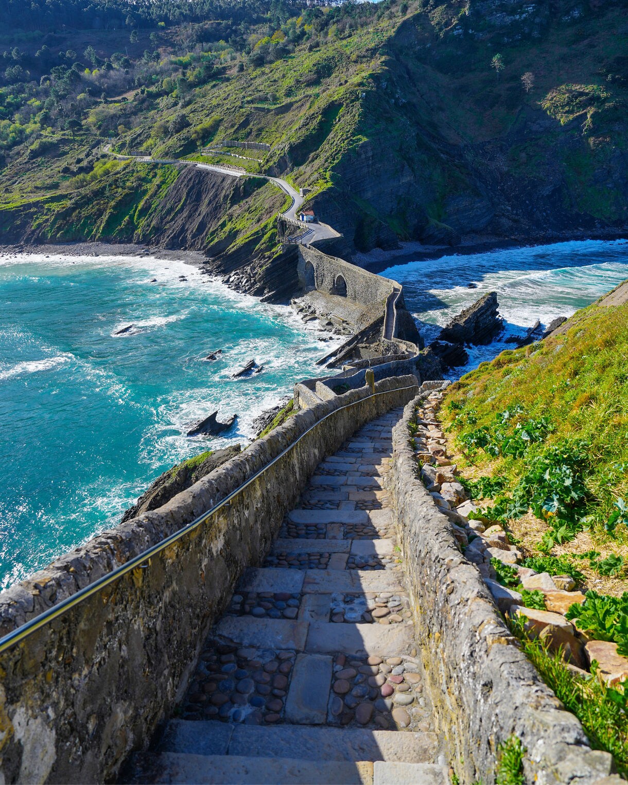 Stone stairway leading down to the rocky path of Gaztelugatxe island in Spain, bordered by turquoise waves and green cliffs under a bright blue sky.